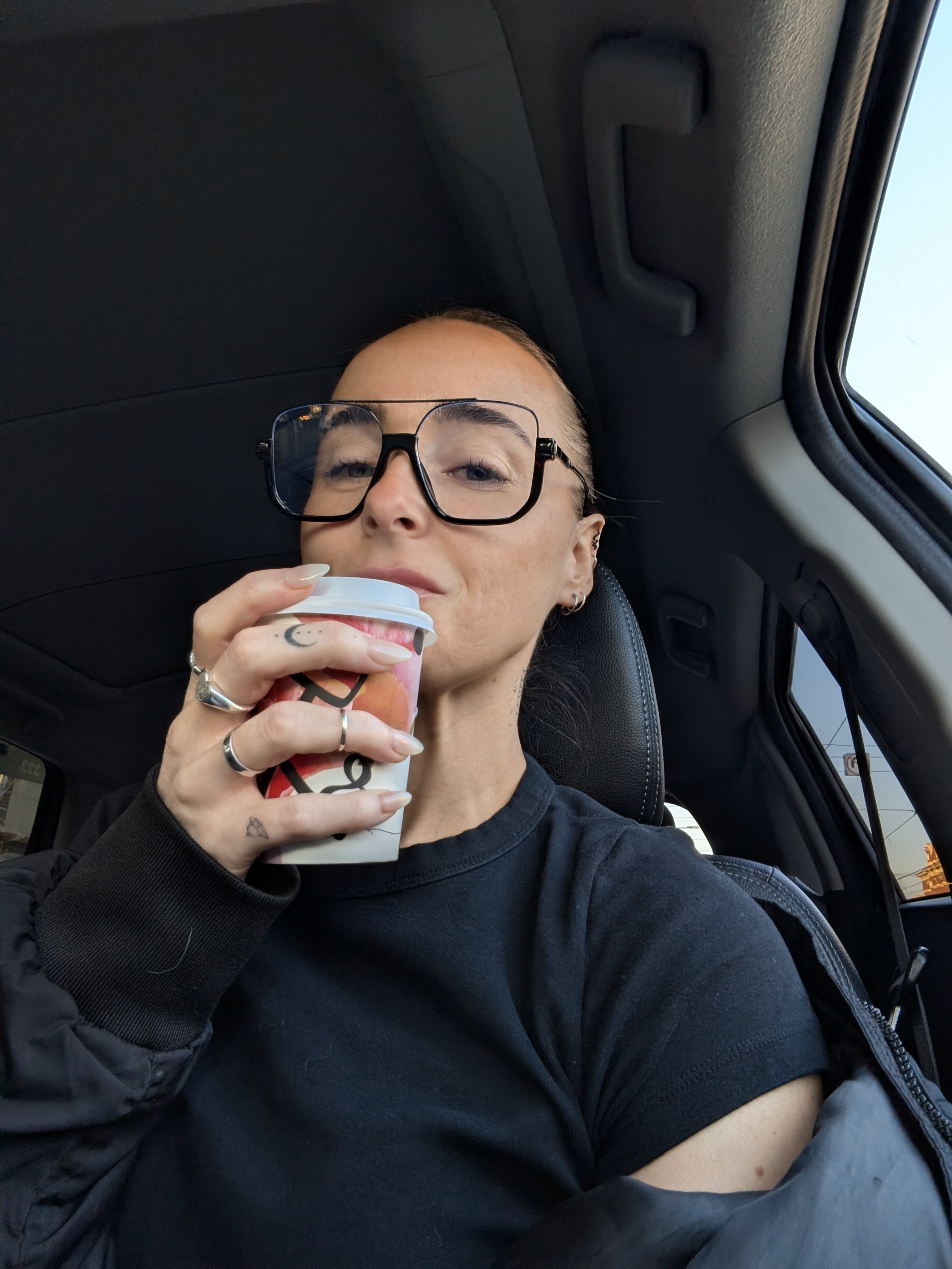 Liv in the driver's seat with oversized black glasses, sipping a takeaway coffee.