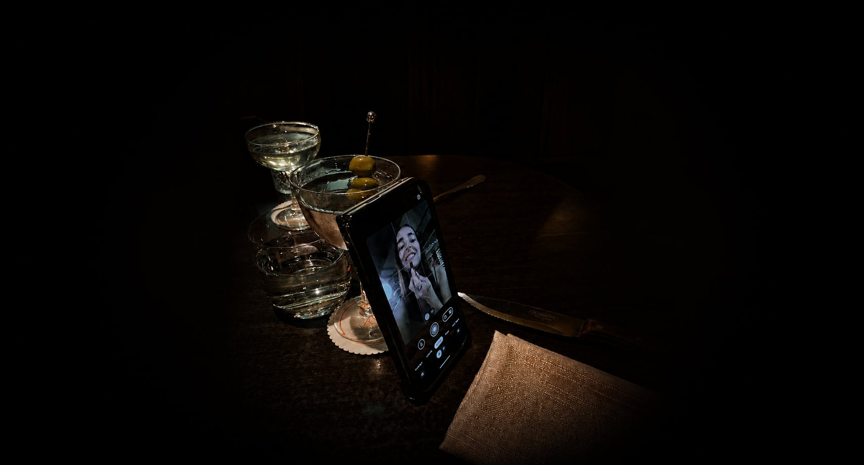 Liv's phone propped upright on a low-lit table beside two martinis, late in the evening.